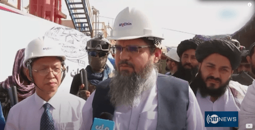 A screen grab of an Ariana News video showing Baradar giving an interview on-site at Kashkari oil field along with high-level Taliban officials. A Taliban guard in military gear can be observed to the left along with a Chinese engineer. The red-colored ba